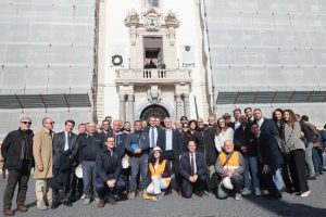 È stata svelata in piazza Duomo a Catania il prospetto restaurato della facciata centrale di Palazzo degli Elefanti, riportando alla luce il volto autentico dell'edificio simbolo della città. L'intervento di tutela e riqualificazione, avviato nel giugno dello scorso anno, è stato finanziato dalla Regione Siciliana e progettato dalla direzione Lavori pubblici comunale. Il sindaco Enrico Trantino ha sottolineato come il `volto´ del Municipio sia il risultato di una ricostruzione filologica condotta in coerenza con le indicazioni della Soprintendenza, evidenziando il ruolo decisivo della ricomposizione cromatica della facciata. Le prove preliminari di pulitura hanno infatti rivelato l'originaria dialettica tra la tonalità giallo sabbia degli intonaci e il candore della pietra calcarea, elementi che nel progetto settecentesco definivano l'equilibrio prospettico del Palazzo degli Eelefanti. Grazie a leggere velature di pigmenti naturali e al ripristino delle finiture originarie, il prospetto riacquista oggi la brillantezza perduta, mettendo in evidenza il ritmo dei pilastri, dei timpani e del grande balcone centrale retto dalle colonne in granito. L'effetto restituito alla piazza richiama l'antica scena urbana immaginata da Vaccarini, in cui la facciata del municipio torna a dialogare con la Cattedrale, il Seminario dei Chierici e il monumento al Liotru, riportando le cromie che per secoli hanno caratterizzato il cuore barocco della città e restituendo ai catanesi un'immagine nitida e fedele del loro palazzo civico. Il restauro ha riguardato soprattutto il consolidamento sismico dell'edificio, cuore istituzionale della città e struttura strategica in caso di emergenza. Costruito nel Settecento sulle rovine della loggia medievale distrutta dal terremoto del 1693, il palazzo chiedeva un intervento urgente finalizzato a preservare la continuità delle murature, spesso indebolite da vuoti interni e discontinuità tipiche dei materiali antichi, affrontato mediante potenti iniezioni di calce fluida resistente per il consolidamento dei paramenti e della copertura in calcestruzzo. Parallelamente al miglioramento strutturale, si è operato sul vasto apparato decorativo, tratto distintivo dell'architettura vaccariniana. Ornamenti e lastre rocciose sedimentate, in parte lesionate o distaccate, sono stati ancorati con barre in vetroresina ad alta aderenza o sostituiti con pietra compatibile con l'originale. Le superfici, offuscate da vecchie tinte e depositi legati al traffico urbano, sono state sottoposte a un accurato ciclo di pulitura che ha rimosso gli strati incoerenti senza intaccare la materia. Sul basamento in pietra lavica si è proceduto con sabbiatura controllata, mentre sulle parti in finta pietra l'integrazione è avvenuta con malte idrauliche pigmentate.